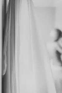 Nahaufnahme eines hängenden Hochzeitskleides aus fließendem Stoff, im Hintergrund unscharf eine Braut beim Getting Ready – elegante Hochzeitsvorbereitungen.