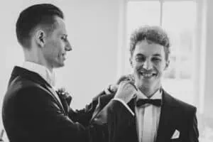 Schwarz-Weiß-Aufnahme eines Bräutigams, der dem Trauzeugen die Fliege richtet, beide lächeln entspannt.