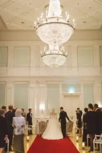 Hochzeitspaar steht im prunkvollen Trausaal des Grand Hotel Heiligendamm unter einem riesigen Kristalllüster, Gäste blicken andächtig auf das Paar.