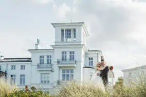 Frisch verheiratetes Brautpaar posiert lachend vor dem Grand Hotel Heiligendamm, umgeben von Dünen und blauer Himmel.