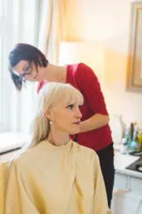 Braut beim Getting Ready: Friseurin stylt die Haare einer blonden Frau am Hochzeitsmorgen in natürlichem Licht.
