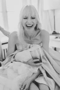 Lachende Braut in Schwarzweißaufnahme beim Getting Ready mit Trinkglas in der Hand – Moment der Freude vor der Hochzeit.