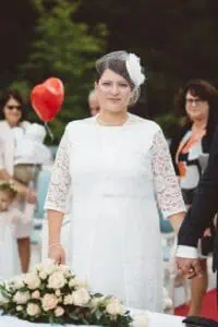 Braut in weißem Spitzenkleid mit Birdcage-Schleier während der Trauung im Grand Hotel Heiligendamm, hält die Hand ihres Partners, im Hintergrund Gäste mit rotem Herzballon.