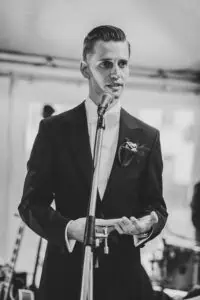 Nahaufnahme eines Bräutigams im eleganten Smoking mit Ansteckblume, wie er während seiner Hochzeitsrede konzentriert ins Mikrofon spricht – Schwarz-Weiß-Fotografie mit emotionaler Wirkung.