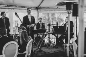 Ein Bräutigam spricht lächelnd ins Mikrofon während seiner Hochzeitsrede, begleitet von einer Live-Band mit Keyboard, Schlagzeug, Gitarre und Saxofon – festliche Atmosphäre in einem Zelt, dargestellt in Schwarz-Weiß.