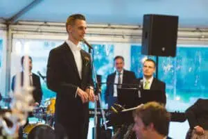 Ein Bräutigam im eleganten Smoking hält eine emotionale Rede bei einer Hochzeit, während eine Live-Band im Hintergrund spielt. Die Atmosphäre ist festlich und musikalisch, mit aufmerksam lauschenden Musikern und Gästen im Bild. Ideal für Inhalte rund um Hochzeitsfotografie, Eventplanung und Livemusik.