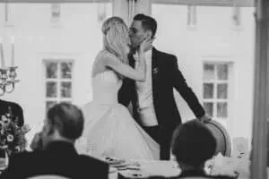 Braut und Bräutigam küssen sich während der Hochzeitsfeier innig an der festlich gedeckten Tafel, Gäste im Vordergrund beobachten das romantische Geschehen.