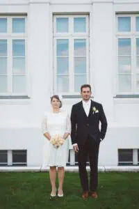 Brautpaar steht auf dem Rasen vor der weißen Fassade des Grand Hotel Heiligendamm, die Braut mit Blumenstrauß, der Bräutigam im Anzug.