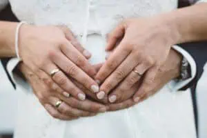 Detailaufnahme der Hände eines Brautpaares mit goldenen Eheringen, liebevoll ineinandergelegt vor weißem Brautkleid, symbolisiert