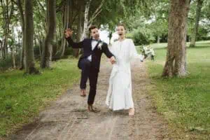 Fröhliches Brautpaar läuft lachend einen Waldweg entlang beim Grand Hotel Heiligendamm, Hand in Hand, voller Lebensfreude.