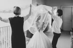 Braut in einem eleganten Hochzeitskleid im Grand Hotel Heiligendamm, unterstützt von zwei Frauen, die ihren langen, verzierten Schleier drapieren.