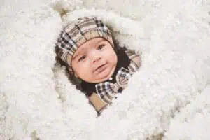 Ein süßes Baby mit einer karierten Mütze und passender Fliege liegt in eine flauschige, cremefarbene Decke eingekuschelt und schaut neugierig in die Kamera.