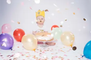 Ein fröhliches Kleinkind mit einer goldenen Krone und der Zahl „1“ sitzt umgeben von bunten Luftballons und Konfetti vor einer Geburtstagstorte. Das Gesicht ist mit Kuchen verschmiert, während es neugierig nach oben schaut.