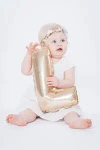 Ein blondes Kleinkind mit blauen Augen und einem Blumenhaarkranz sitzt barfuß auf einem weißen Untergrund und hält einen goldenen Folienballon in Form der Zahl „1“ fest.