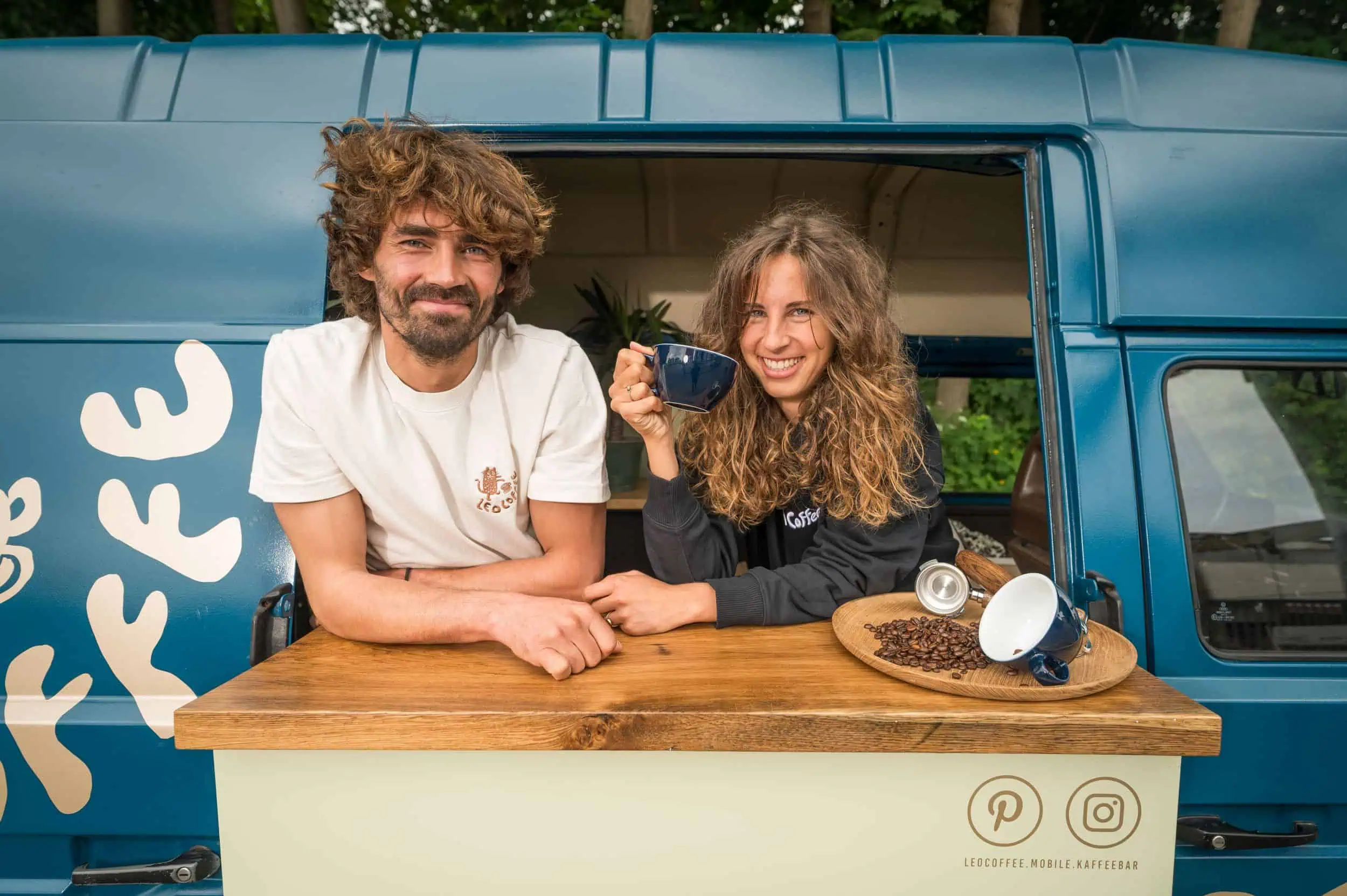 Rostocker gründen mobilen Kaffee-Bulli Ronja Arndt und Johannes Witte an ihrer mobilen Kaffeebar von Leo Coffee vor einem blauen Kaffeewagen, mit Espressotasse und Kaffeebohnen.