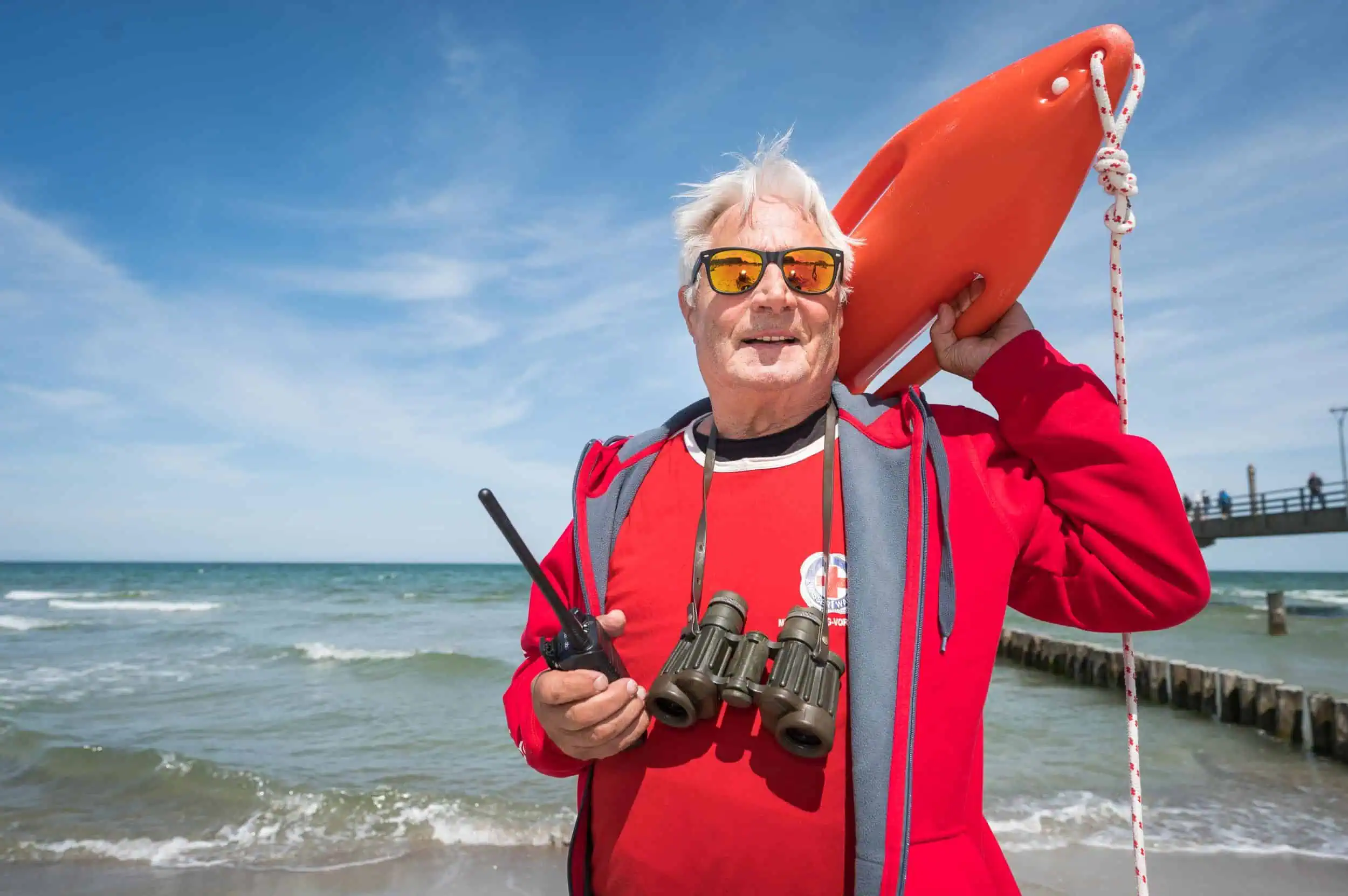 Ein ältere Rettungsschwimmer in posiert für den Fotografen Lothar Kock, älterer Rettungsschwimmer mit roter Kleidung und Sonnenbrille steht am Strand, hält eine Rettungsboje und ein Funkgerät, mit dem Meer und einer Seebrücke im Hintergrund.