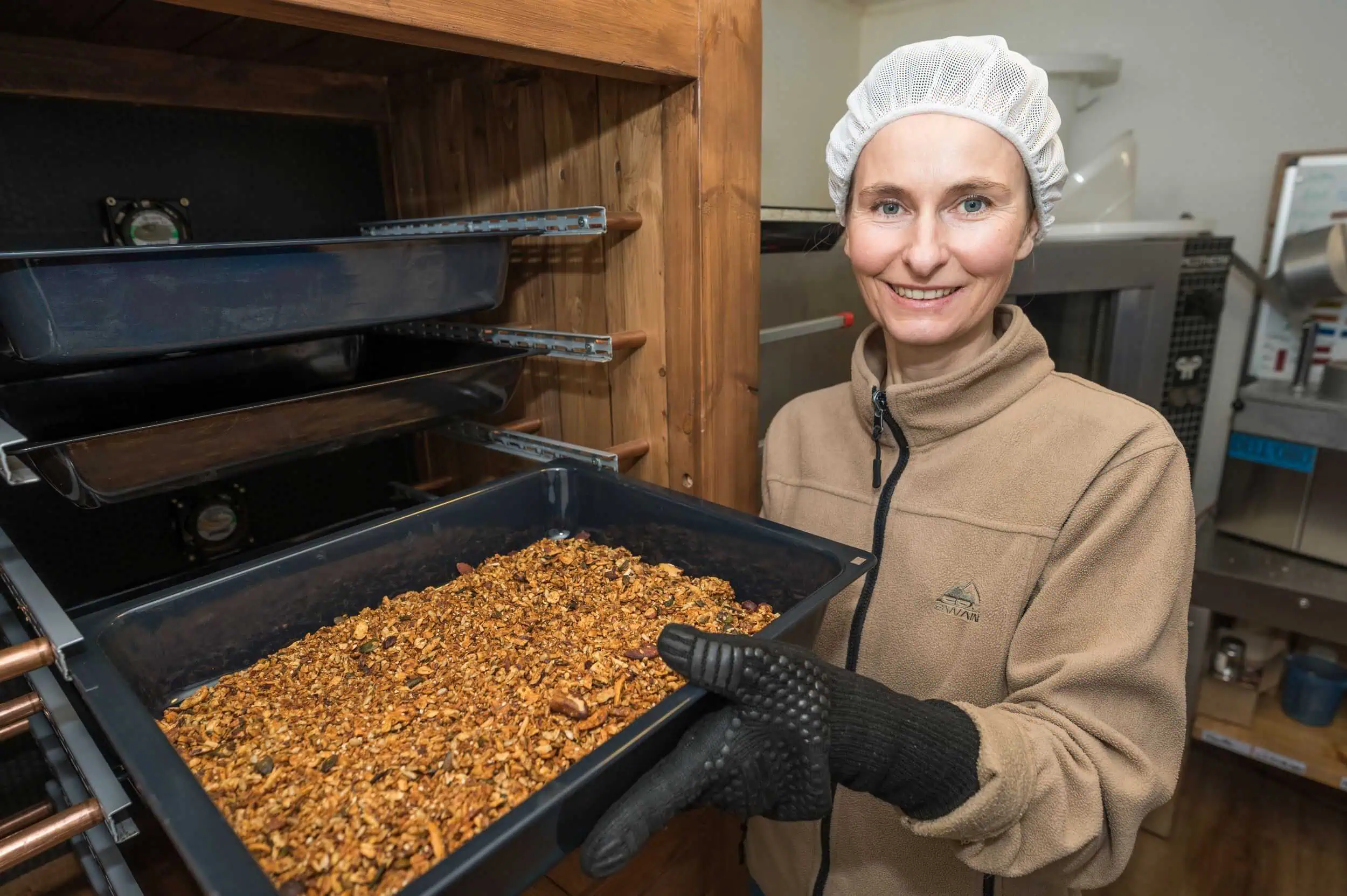 Nööt Tied Janine Kohler-Seupel mit Haarnetz und Handschuhen hält Blech mit knusprigem Granola vor einem Ofen.
