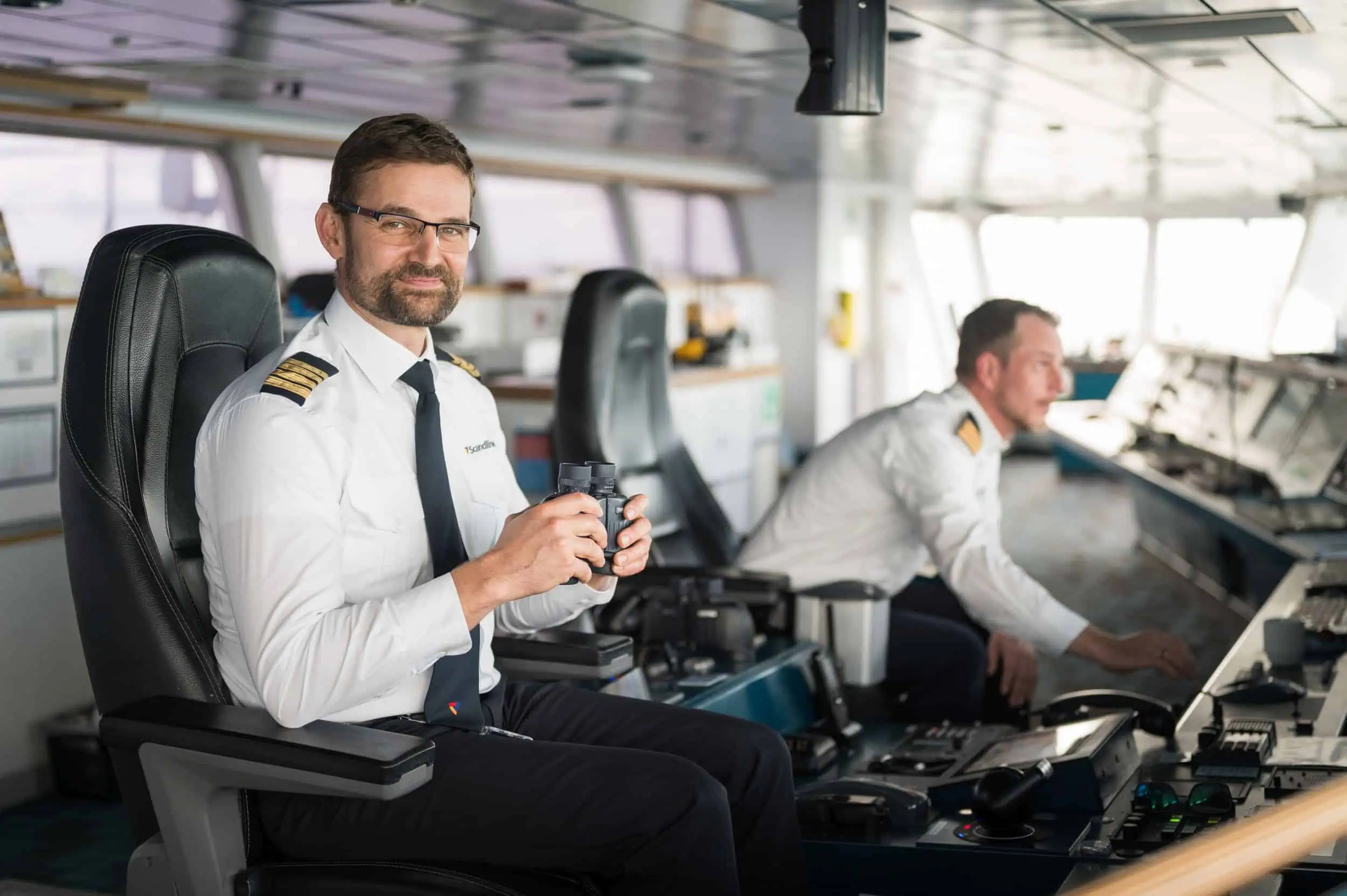 Kapitän Stefan Wehland Kapitän Stefan Wehland auf der Brücke einer Scandlines Fähre mit Fernglas in der Hand, im Hintergrund ein weiterer Offizier am Navigationspult.