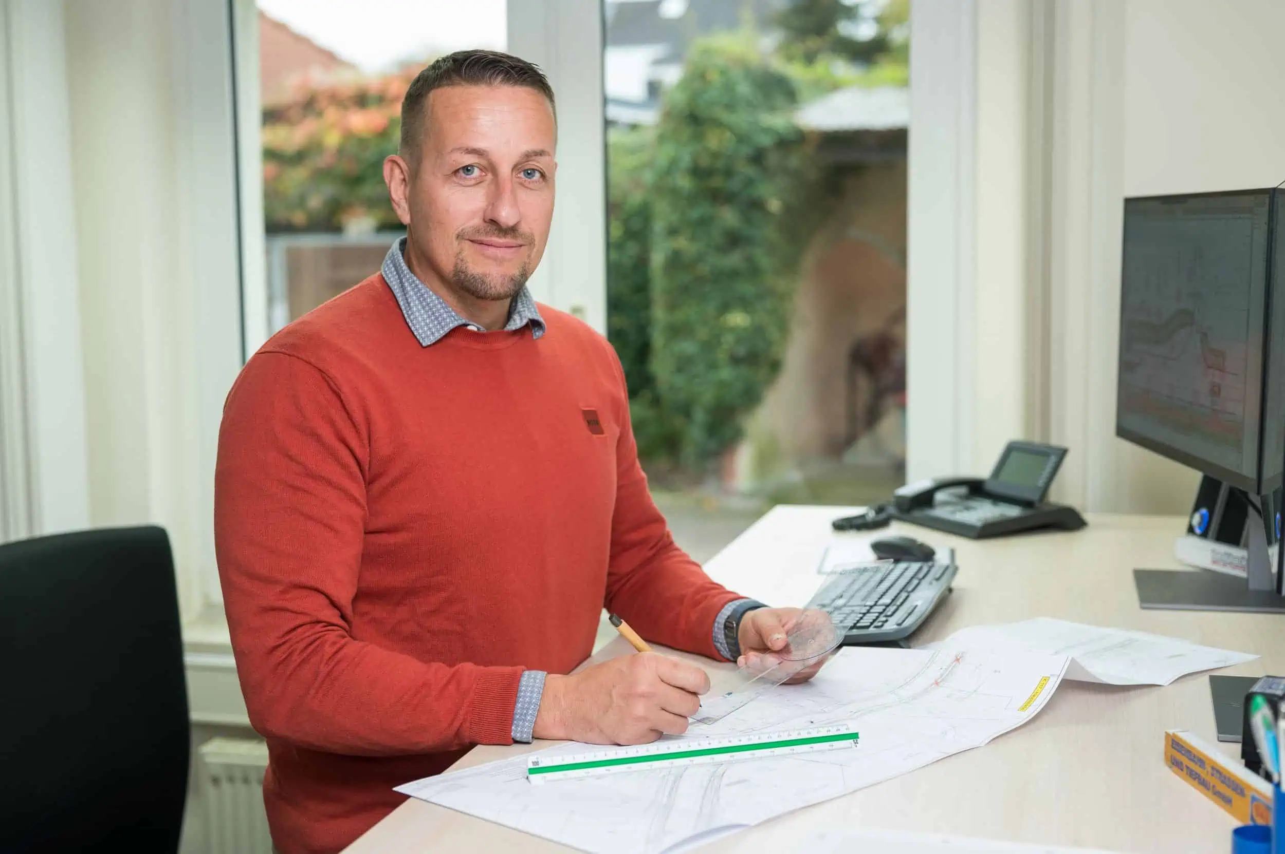 Martin Boerner Mitarbeiter mit rotem Pullover arbeitet am Schreibtisch an Bauplänen mit Lineal und Computer.
