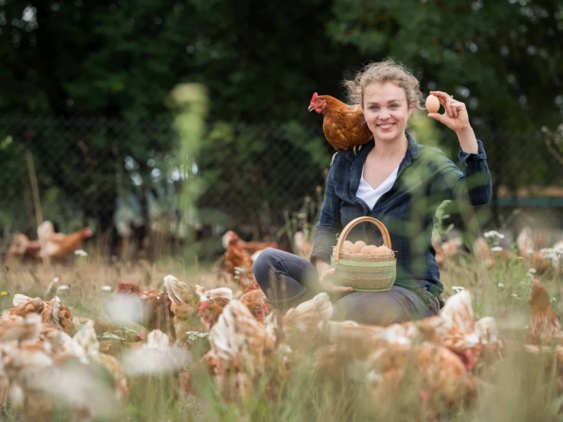 Eier Abo aus MV Leonie Behrens sitzt inmitten einer Hühnerschar auf der Wiese mit einem Korb voller Eier und einem Huhn auf der Schulter.
