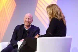 Matthias Sammer sitzt auf einer Bühne bei einer Veranstaltung in Hohe Düne im Gespräch mit einer Moderatorin. Er trägt ein dunkles Sakko und lächelt, ein Headset-Mikrofon ist sichtbar. Im Hintergrund fallen farbige Lichtreflexe auf die Wand. Foto: Martin Börner, Eventfotograf aus Rostock.