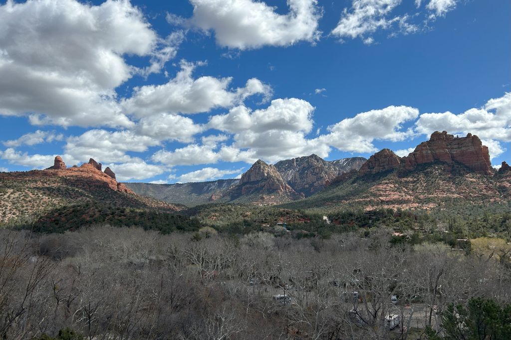 Sedona Az Spiritual Place (2026 Local Guide) This photograph showcases a breathtaking view of the Sedona landscape in Arizona. It features a panorama of red rock formations including Bell Rock on the left, distinctive for its smooth, bell-like shape, and the layered cliffs of Courthouse Butte to the right. The scenery is set against a dynamic sky filled with puffy clouds, and the valley below is dotted with leafless trees and several buildings, depicting a serene suburban setting nestled within the natural beauty.