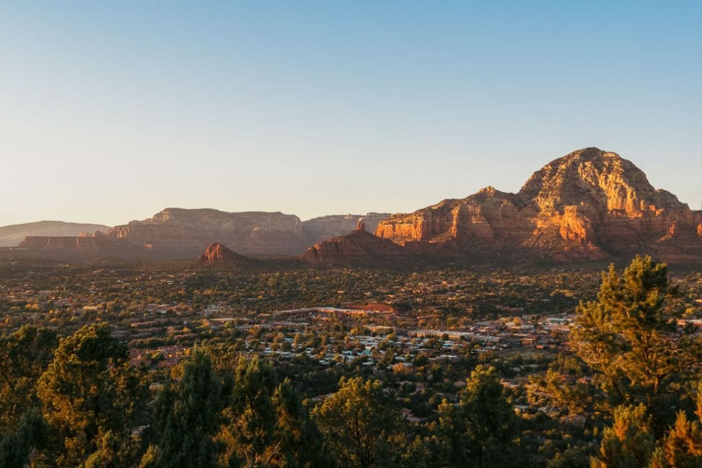 Sedona Az Spiritual Place (2026 Local Guide) This image captures a serene sunset view over Sedona, Arizona, with its iconic red rock formations bathed in the warm glow of the setting sun. The expansive view highlights the town nestled among the majestic landscape, surrounded by a panorama of rugged cliffs and plateaus. The clear sky and the golden sunlight enhance the rich, natural colors of the area, providing a picturesque scene typical of the American Southwest.