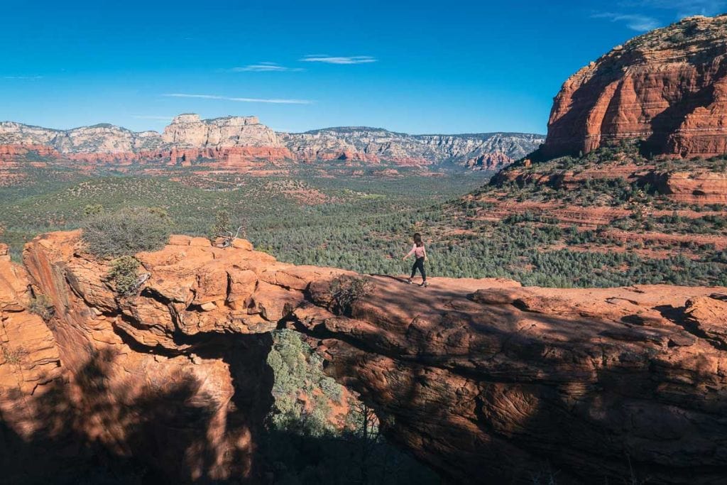 Sedona Az Spiritual Place (2026 Local Guide) This image shows a breathtaking view of Devil's Bridge in Sedona, with a person cautiously walking across the natural red sandstone arch. The arch overlooks a vast landscape of lush greenery and layered red rock formations, under a clear blue sky. The scene captures the adventurous spirit and stunning geological beauty typical of Sedona's rugged terrain.