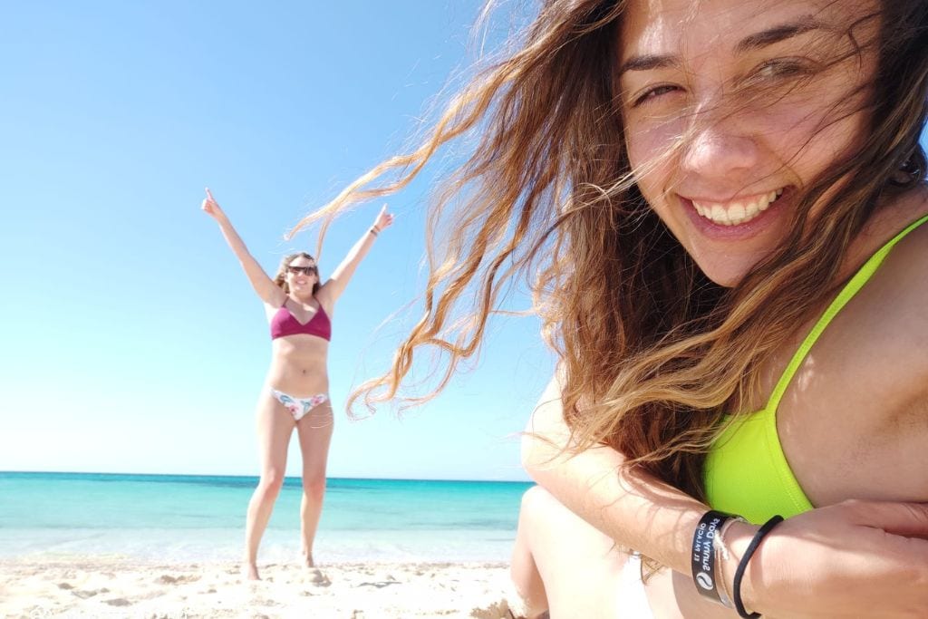 Two women, aka me and Lea, are joyfully posing on a sunny beach at the red sea; one is in the foreground taking a selfie, her hair swept by the wind, and the other stands in the background with her arms raised in a carefree gesture. The turquoise sea behind them reflects the clear sky, creating a vibrant and happy seaside snapshot.