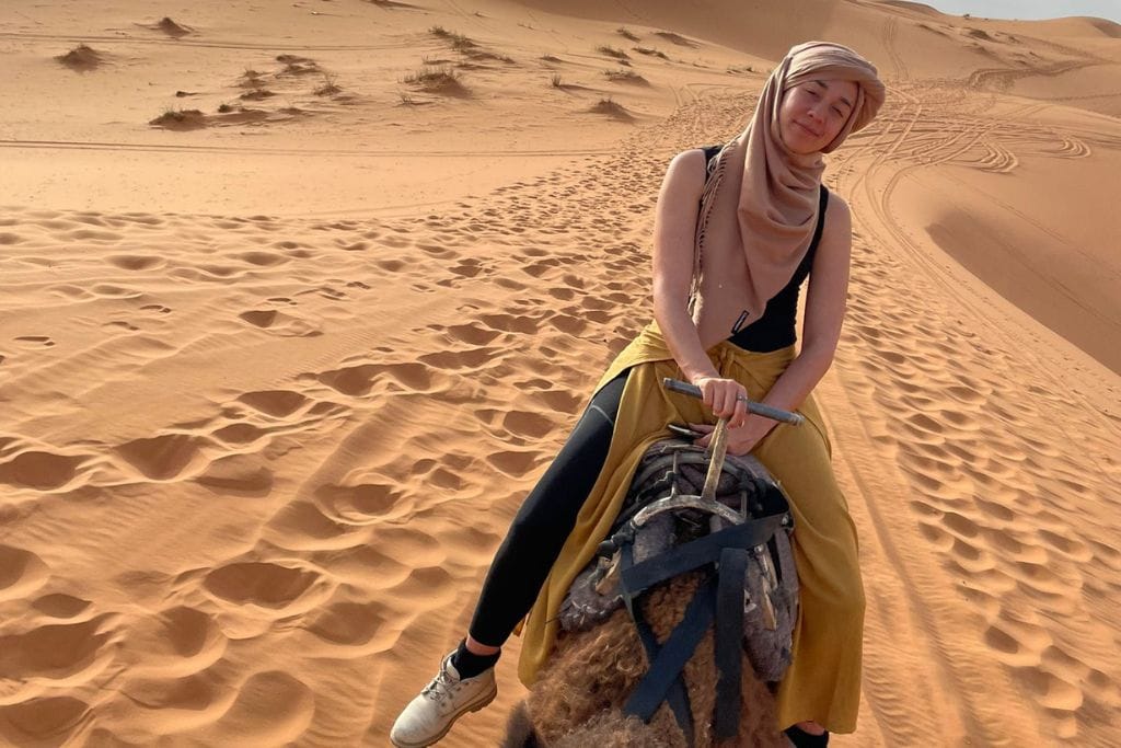 A smiling woman, wrapped in a headscarf, sits atop a camel in the desert, with rolling dunes in the background and the impressions of many footsteps in the sand around her.