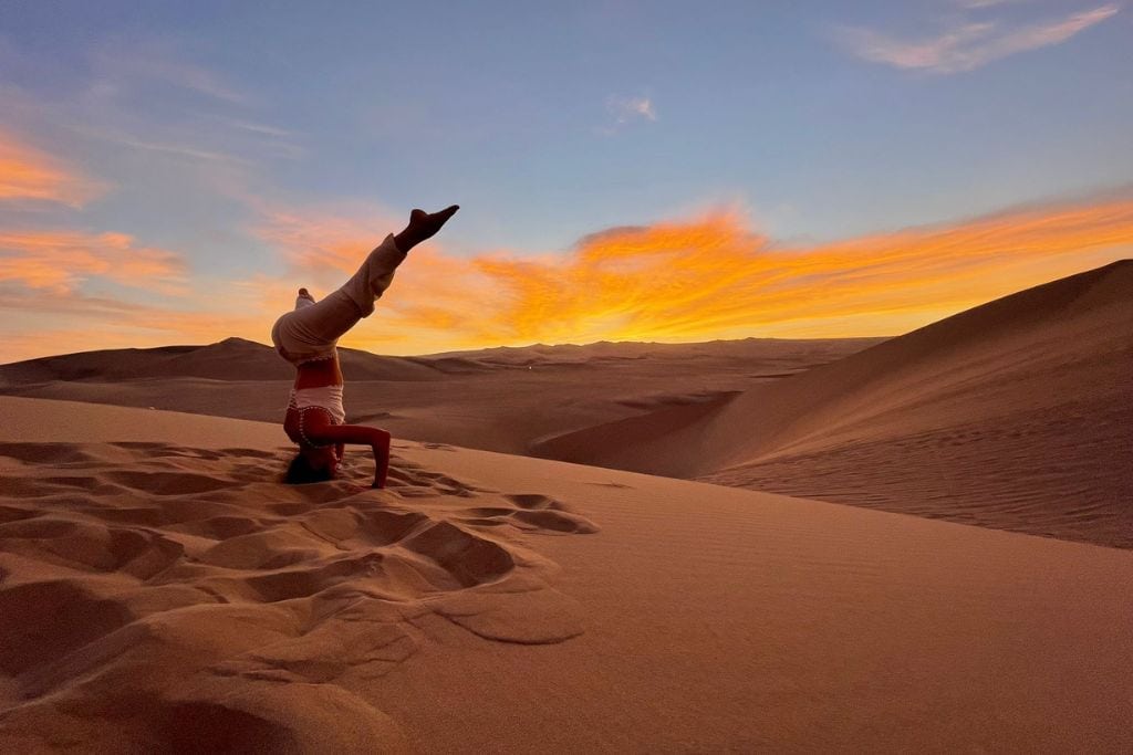 A person performs a headstand on the Sahara desert dune against a vibrant sunset with streaks of orange and pink in the sky. Why is the Sahara Desert Spiritual Awakening?