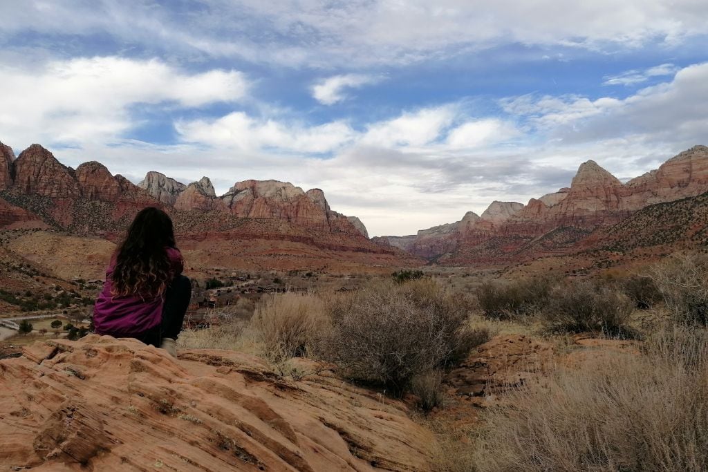 A person sits contemplatively on the red rocks of Sedona, AZ, overlooking a scenic view of rugged mountains, under a vast sky, embodying the tranquility and spiritual essence of this majestic place.