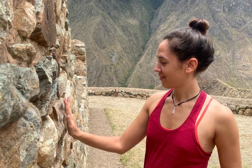 A woman in a red tank top and blue patterned leggings stands touching an ancient stone wall, gazing to the side with a soft smile. The mist-covered Andean mountains provide a dramatic backdrop to this serene moment, capturing a personal connection with the historic Inca Trail in Peru. Way up to the Machu Picchu Spritiual!