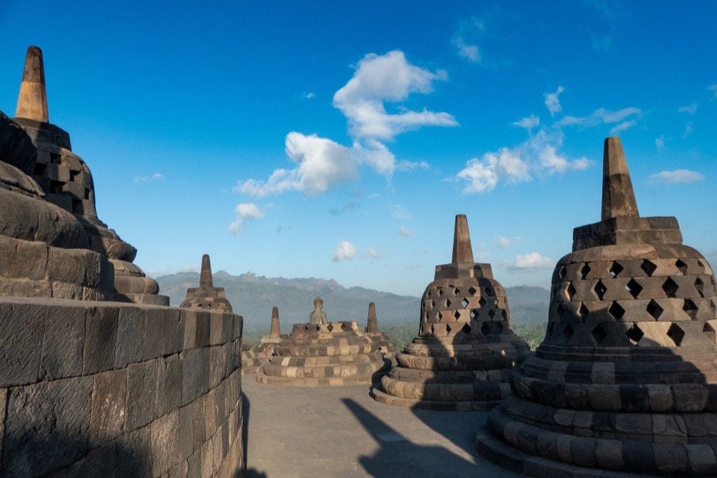 Rows of stone stupas, each with a distinct lattice design, line the upper terraces of Borobudur Temple, converging towards a serene Buddha statue in the center. The clear blue sky above provides a vibrant backdrop to this historic spiritual site.