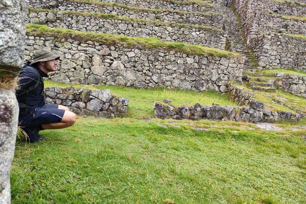 A man in hiking attire squats contemplatively beside ancient stone terraces, his gaze fixed on the green grass that carpets the archaeological site.