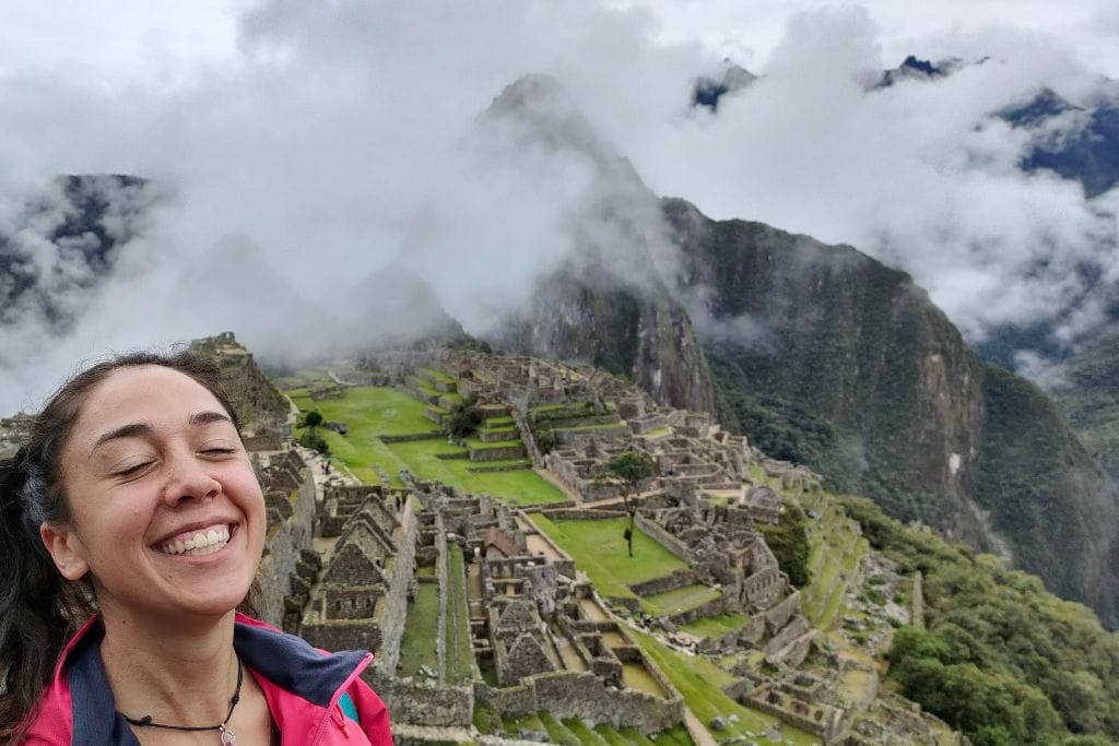 A joyful woman takes a selfie with the iconic Machu Picchu ruins in the background, partially veiled by mist, showcasing the magnificent blend of human history and natural beauty.