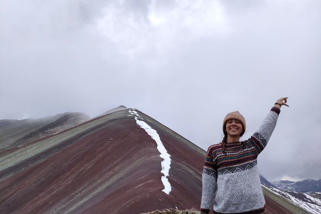 A smiling person in a beanie and sweater pointing towards the sky on the multicolored slopes of Rainbow Mountain, Peru, with a cloudy sky overhead. Rainbow Mountain Peru Spiritual Meaning (Vinicunva Sacred Facts)