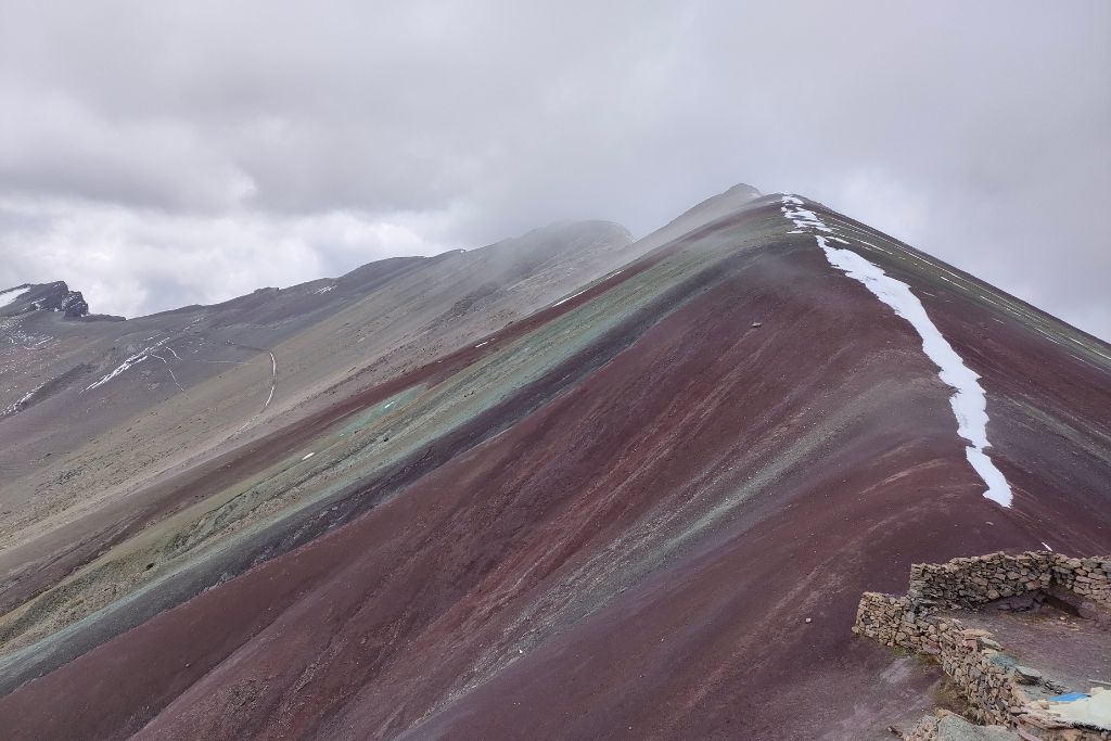 Rainbow Mountain Peru: Your Ultimate Spiritual Guide The striking striped hues of red, turquoise, and lavender along the slope of Rainbow Mountain, Peru, with a cloudy sky overhead and remnants of snow tracing the mountain's contours.