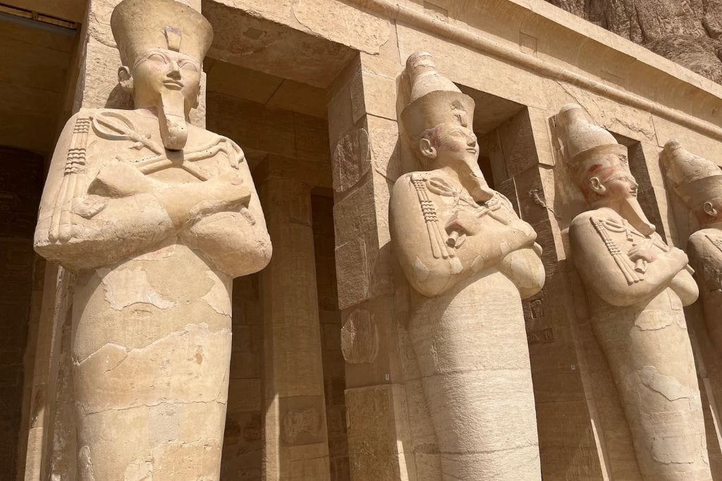 Luxor City in Egypt: Is it Worth Visiting? Close-up view of weathered ancient Egyptian statues with crossed arms, depicting pharaohs wearing traditional headdresses, at the Temple of Hatshepsut in Luxor, with the limestone cliffs of Deir el-Bahari in the background.