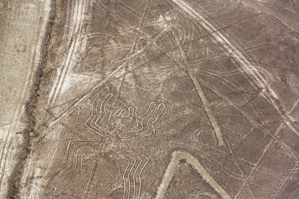 Why are the Lines of Nazca a Mystery of Peru? Aerial view of the spider geoglyph, one of the mysterious Lines of Nazca, with its elongated legs and distinct body etched into the Peruvian desert, surrounded by intersecting lines and geological textures. The Lines of Nazca.