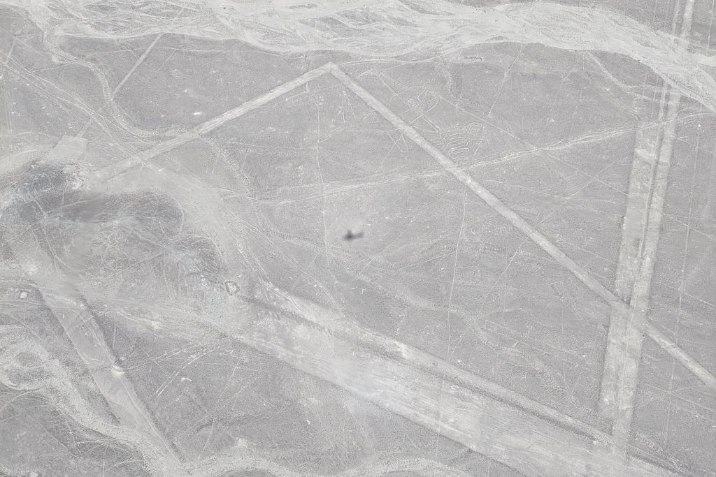 Why are the Lines of Nazca a Mystery of Peru? Overhead view of the faint outlines of the Nazca Lines, including a spiral and a bird-like figure, interwoven with the natural patterns and ridges of the desert landscape in Peru.