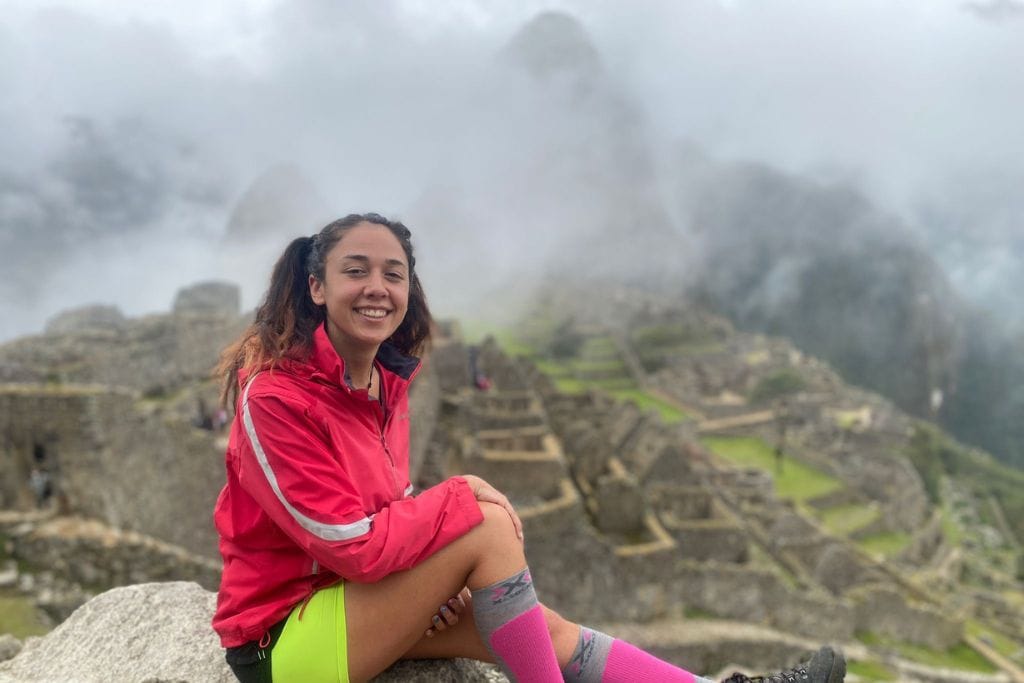 A smiling young woman sits on a rock at Machu Picchu spiritual, with the iconic fog-shrouded ruins in the background. She wears a bright red rain jacket, neon green shorts, and pink socks, complementing the vibrant energy of her adventure in the Peruvian Andes.
