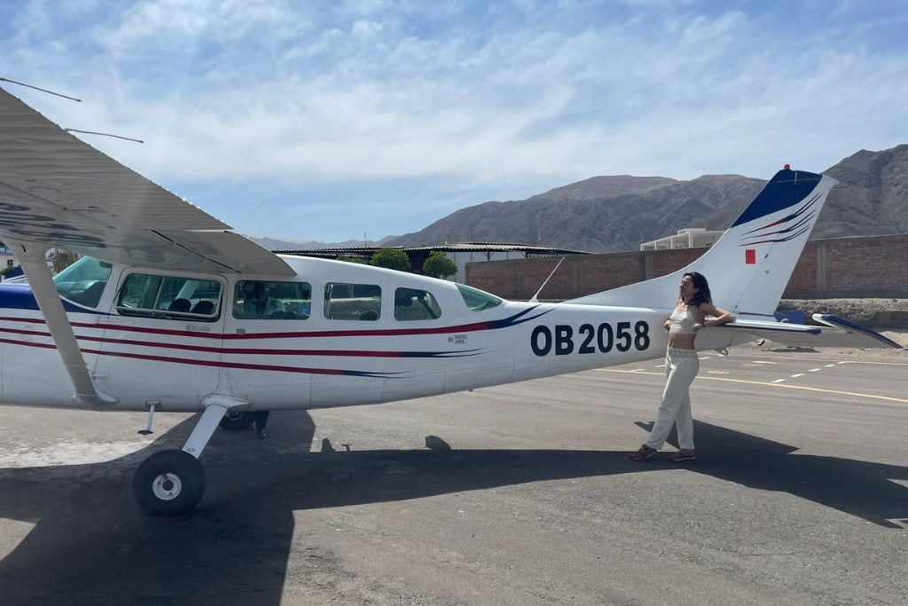Why are the Lines of Nazca a Mystery of Peru? Woman in white clothing posing with her hand on her hip next to a small, white airplane with registration OB2058, ready for flying over the lines of Nazca.
