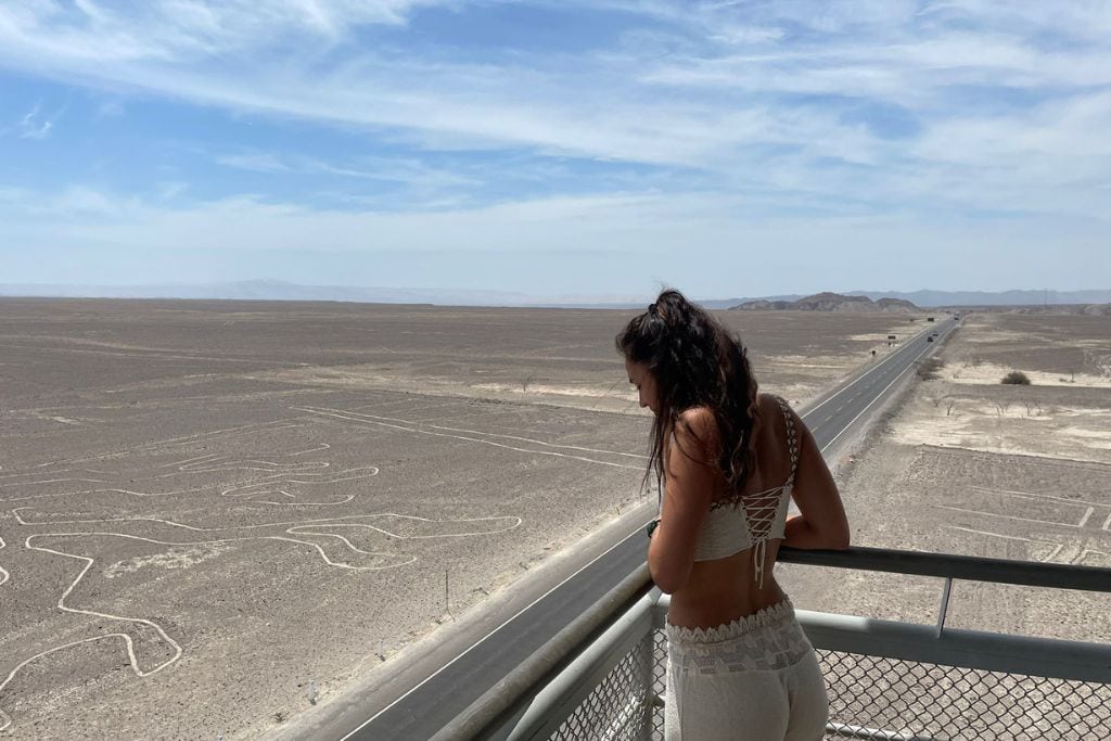 Why are the Lines of Nazca a Mystery of Peru? Woman in a white outfit looking down from an observation deck at the Nazca Lines, with the vast, textured desert expanse and a straight highway stretching into the distance.