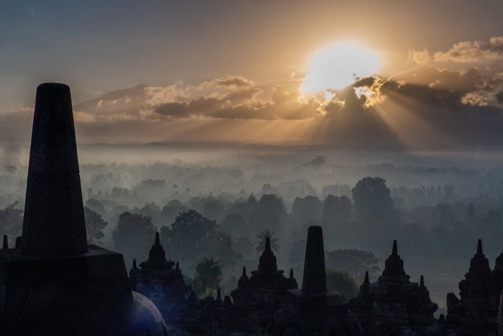 The sun rises over the silhouette of Borobudur Temple, with rays piercing through the misty forest canopy. This serene dawn scene captures the historic stupa-shaped terraces amidst a mystical landscape. Visiting Borobudur