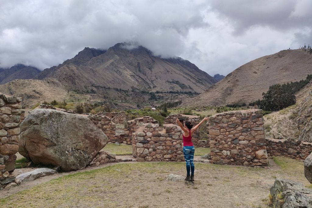 A woman with raised arms stands between two ancient stone walls amidst a rugged mountainous landscape. Overcast skies loom above the peaks, and the sense of history in the air is palpable.
