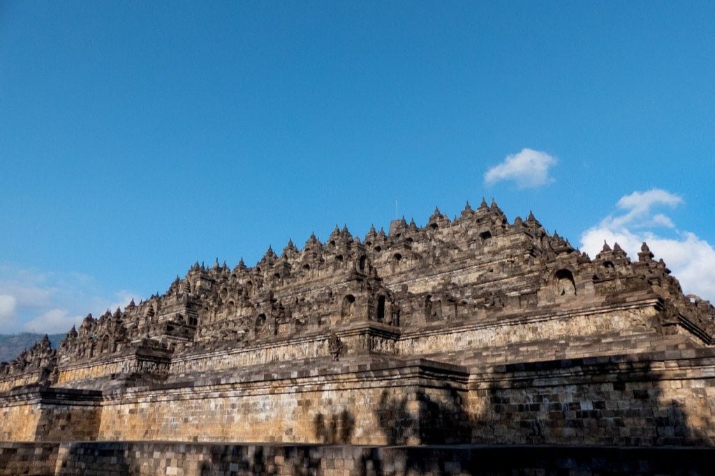 The grandeur of Borobudur Temple's stepped design stands against a clear blue sky. The intricate stone carvings and bell-shaped stupas are a testament to the temple's historical and architectural significance.