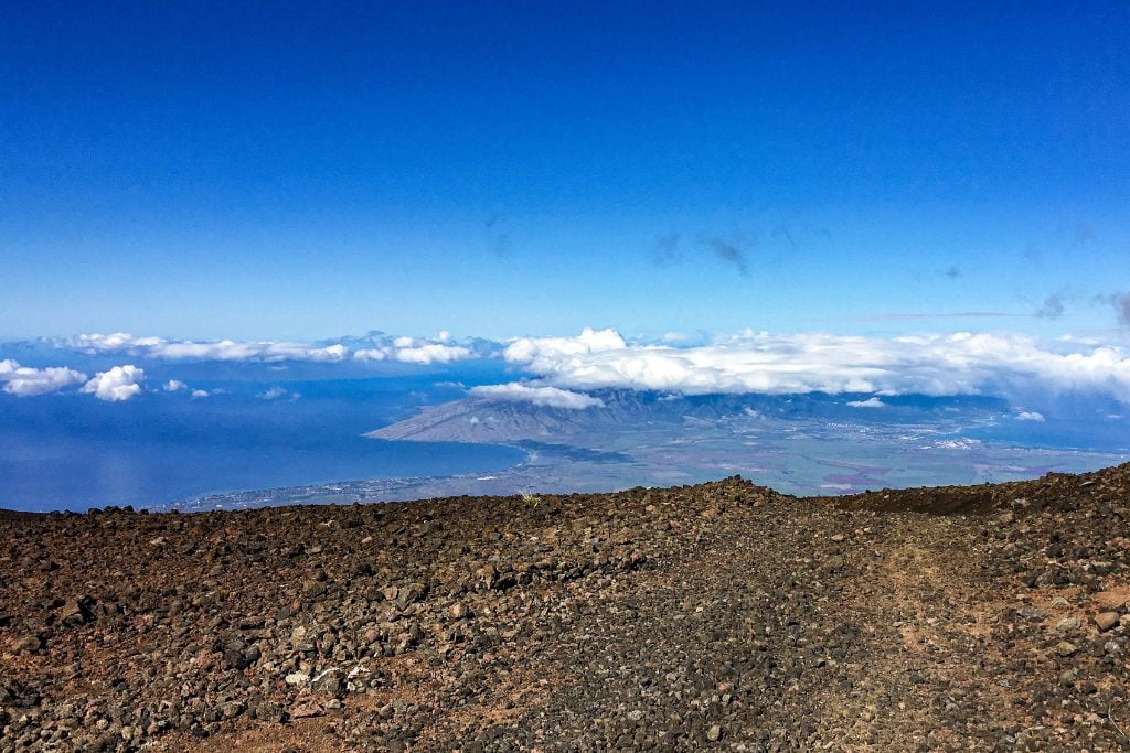 A panoramic view from the summit of Haleakalā, showcasing a vast expanse of rugged volcanic terrain in the foreground and a distant view of Maui's coastline and clouds on the horizon under a clear blue sky. The contrast between the barren lava rock and the lush island below evokes the diverse beauty of Hawaii's landscapes.