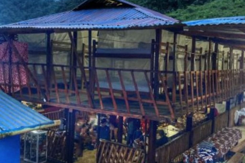 How to Get to the Lost City Colombia? My Personal Experience in 2026! A campsite with bunk beds under a metal roof, surrounded by forested hills, and illuminated in the evening light. The camp features a communal area with benches and tables, and a few people can be seen gathered inside.