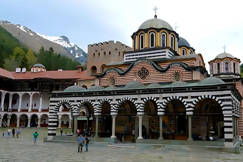 The Rila Monastery in Bulgaria, showcasing its distinctive black-and-white striped arches and domes, set against a backdrop of snow-capped mountains and lush green forests. Visitors can be seen exploring the courtyard of this historic and ornate Eastern Orthodox monastery.